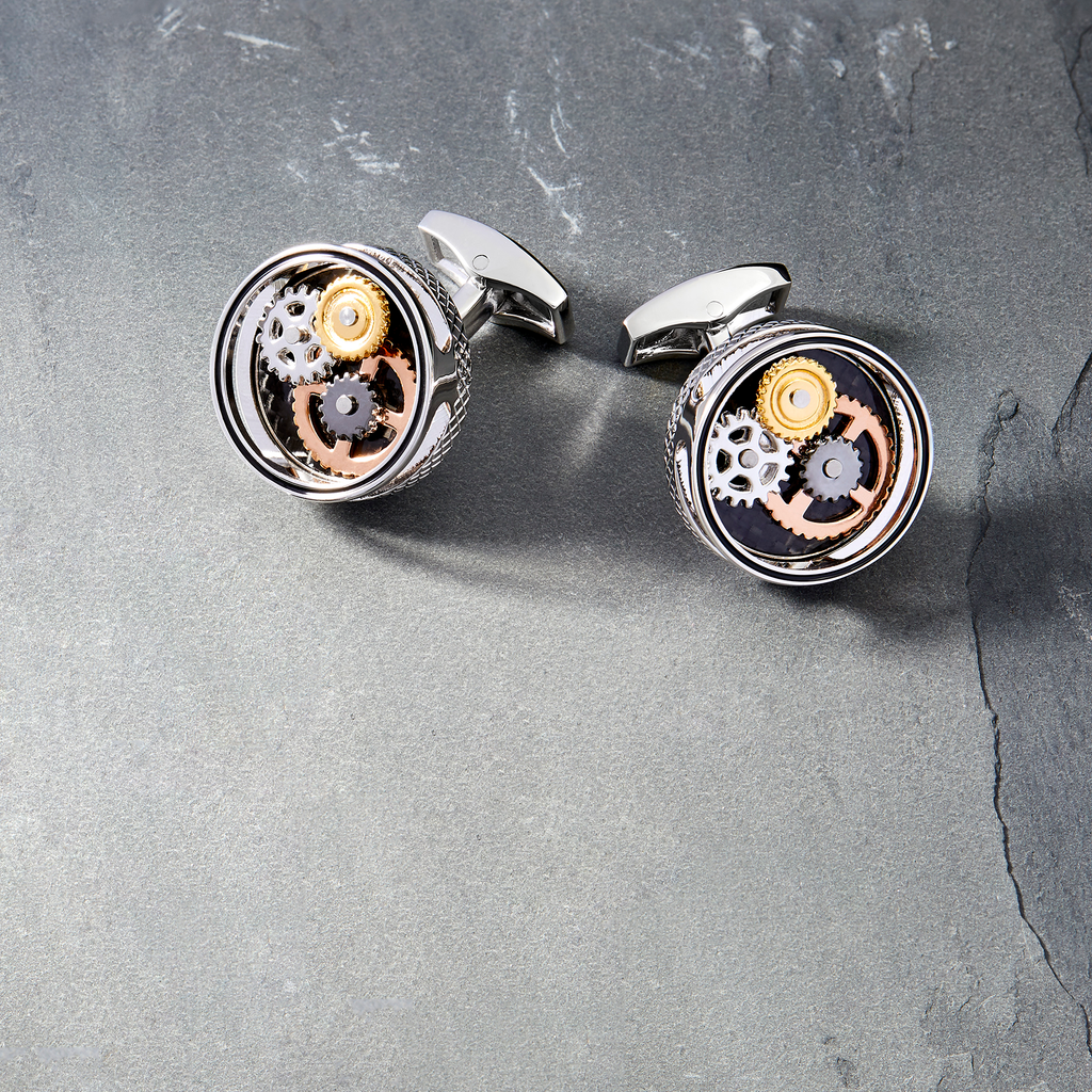 A pair of palladium-plated cufflinks with a round shape featuring rotating gear mechanisms and a black carbon fiber inlay.